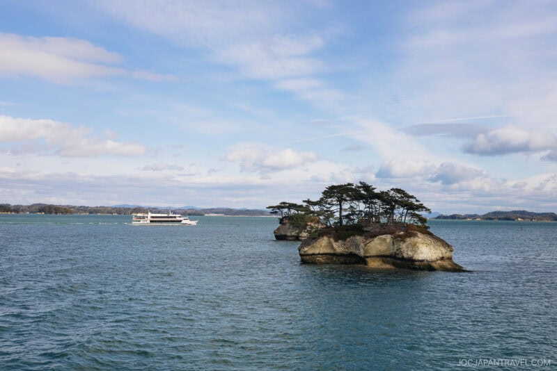 A small rocky island with pine trees sits in blue water under a partly cloudy sky. In the distance, a white sightseeing boat sails across Matsushima Bay.