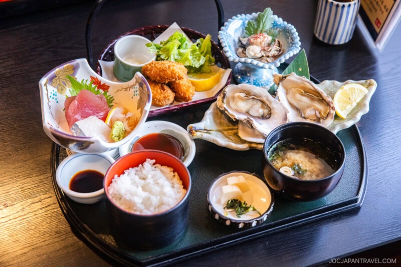 A Japanese seafood meal set on a black tray, featuring fresh sashimi, oysters from Matsushima, fried croquettes, salad, rice, miso soup, dipping sauces, and garnishes.