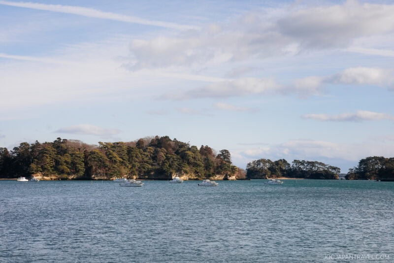 Several small boats float on calm blue water near a tree-covered island under a partly cloudy sky in scenic Matsushima.