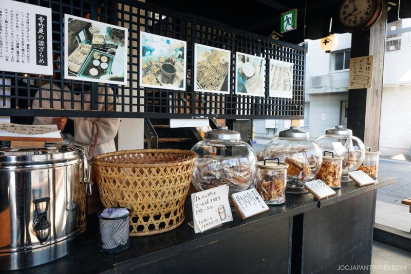 Glass jars filled with traditional Japanese snacks are displayed on a wooden counter in Matsushima, with handwritten labels and photos above describing each item. A woven basket and metal pot are also visible in the inviting setup.