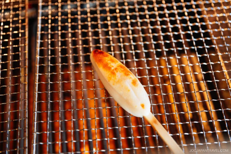A skewered piece of grilled mochi is being roasted over a metal grate with visible flames underneath, capturing the flavors and traditions often found in Matsushima.