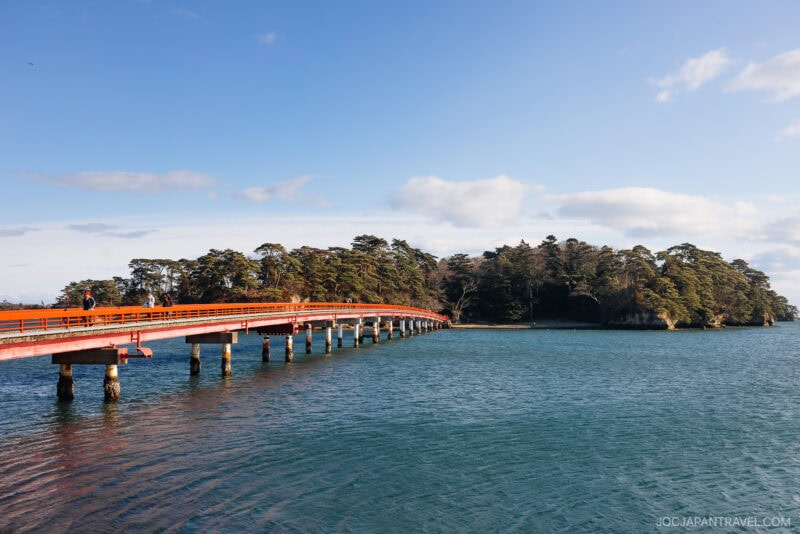 A long red bridge in Matsushima stretches over blue water toward a tree-covered island under a clear sky with scattered clouds.
