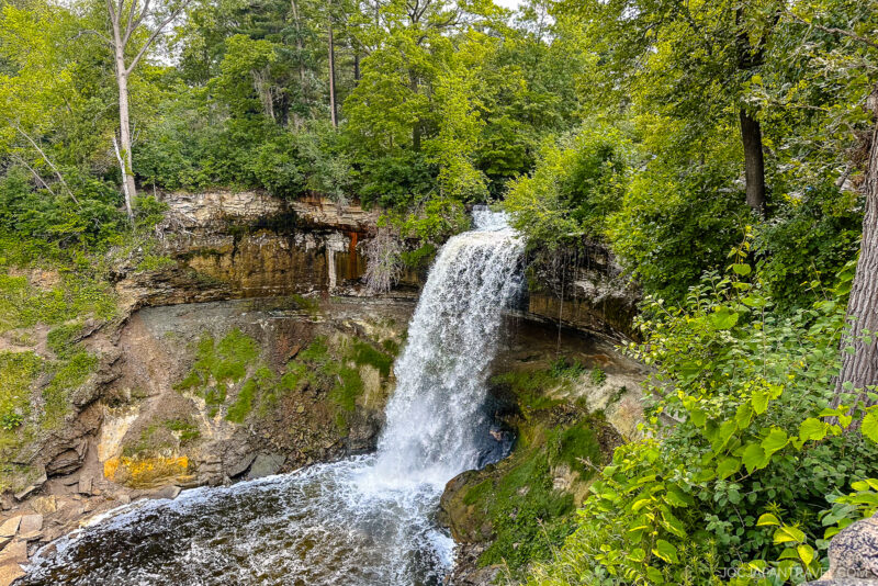 A waterfall cascades over a rocky cliff into a pool below, surrounded by lush green trees and foliage on a bright, sunny day in Minneapolis.