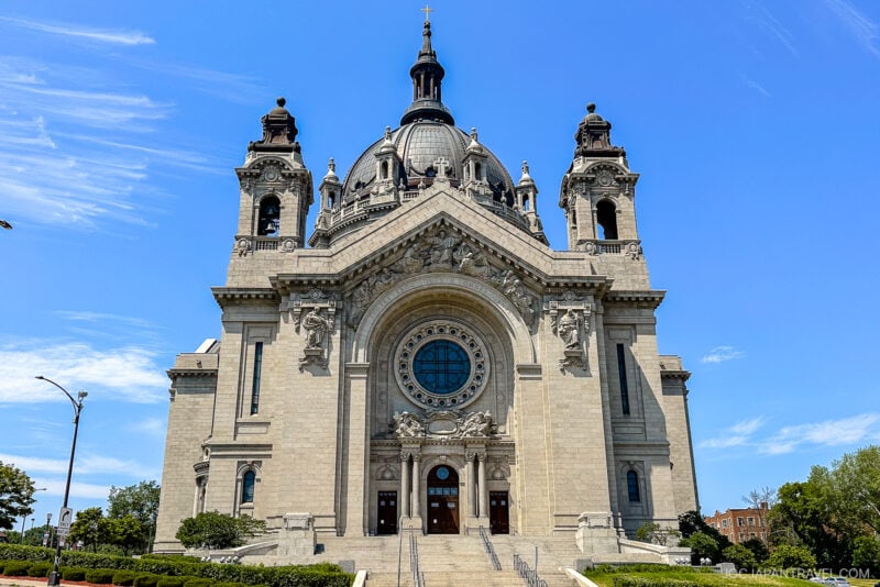 Front view of the grand, ornate Minneapolis cathedral with a large central dome, twin towers, arched entrance, and circular stained-glass window, set against a bright blue sky with wispy clouds.