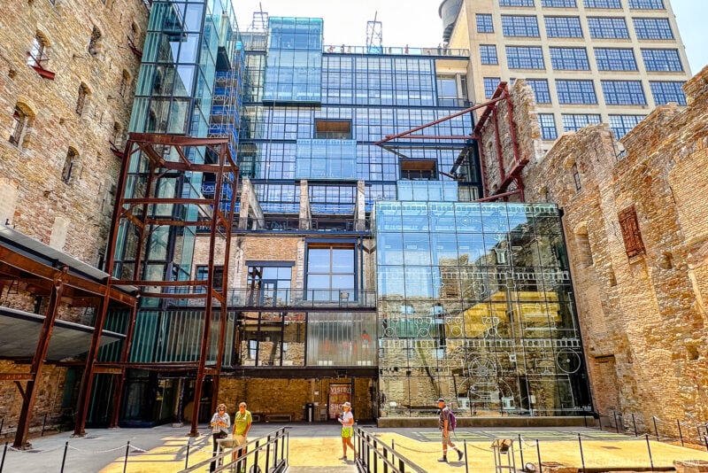 People walk and play in a sunny Minneapolis courtyard surrounded by historic brick ruins and modern glass architecture, blending old and new building styles.