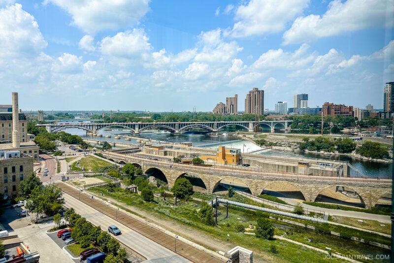 A Minneapolis cityscape with multiple bridges crossing a wide river, surrounded by greenery and mid-rise buildings under a partly cloudy sky. Railroad tracks and roads run alongside the river in the foreground.