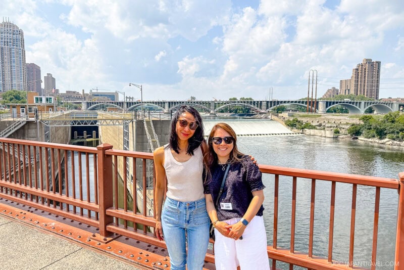 Two women stand smiling by a red railing in front of a river in Minneapolis, with multiple bridges and city buildings in the background on a sunny day.
