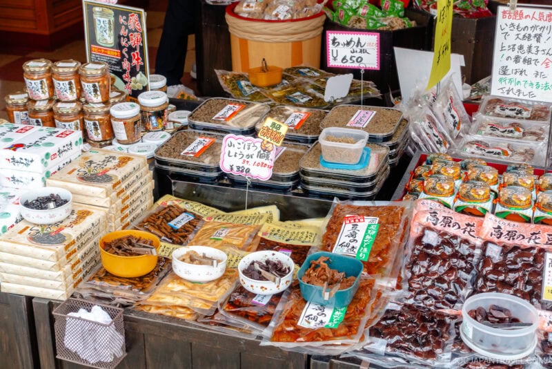 A colorful Miyagawa Morning Market stall displays various packaged Japanese foods, jars, dried goods, and snacks with handwritten Japanese labels and price tags on each item.