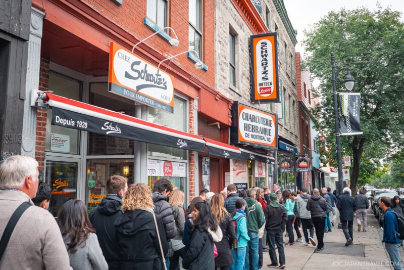A crowd of people lines up outside Schwartz’s Deli, a legendary spot in Montreal food culture, with red brick walls and bold signage on a busy city street. Trees and other storefronts add to the lively scene.
