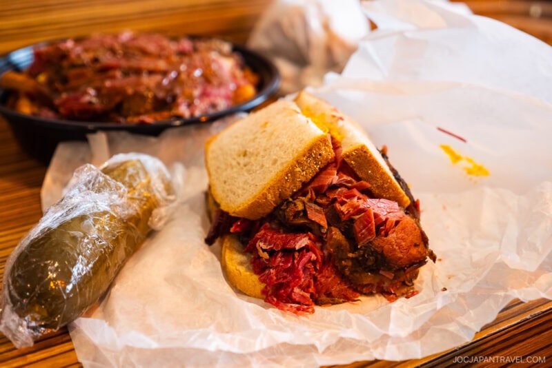 A pastrami sandwich on white bread sits on wax paper next to a whole pickle wrapped in plastic. In the background, a container holds sliced meat—a classic scene of Montreal food, set on a wooden table.