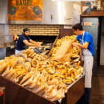 Where to Eat in Montreal: Family-Friendly Food Guide Two workers in a bakery handle large piles of bagels—one fills a paper bag while the other arranges fresh bagels by the brick oven. A sign above reads Bagel Shop Montréal, capturing the spirit of classic Montreal food.