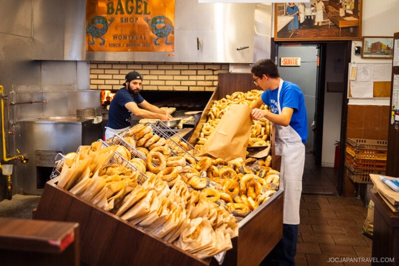 Two workers in a bakery handle large piles of bagels&mdash;one fills a paper bag while the other arranges fresh bagels by the brick oven. A sign above reads Bagel Shop Montr&eacute;al, capturing the spirit of classic Montreal food.