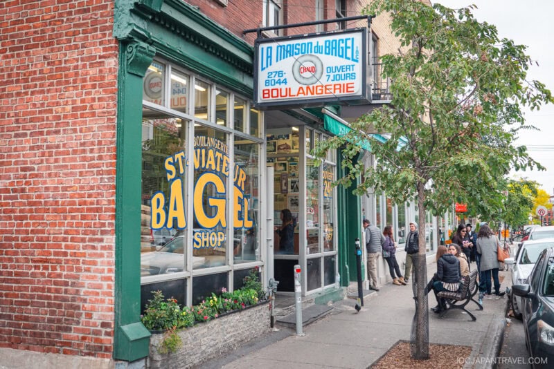 Street view of St-Viateur Bagel shop in Montreal, a staple of Montreal food, with a large yellow and blue sign on the window, people lined up outside, and a tree along the sidewalk in front of the brick building.