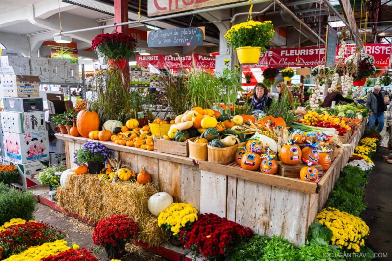 A vibrant farmers market stall in Montreal displays pumpkins, gourds, squash, and colorful flowers. Signs read Fleuriste above the stand, celebrating local Montreal food as shoppers browse in the background under a covered roof.