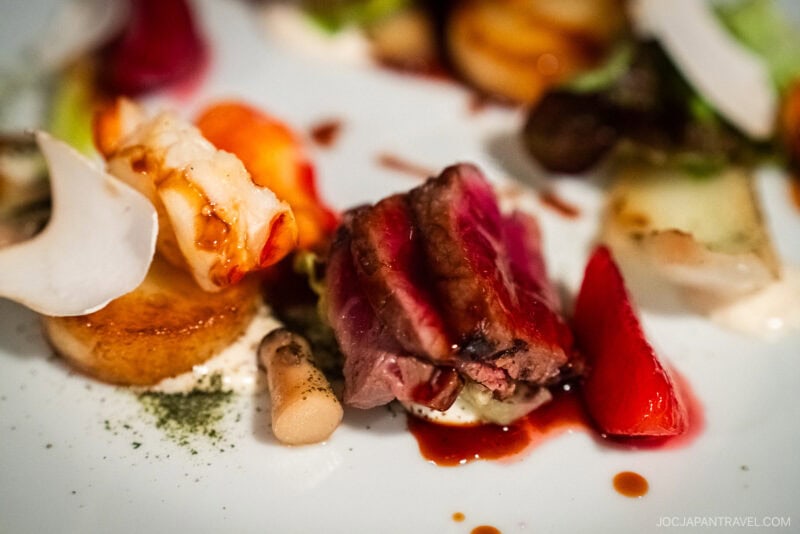 A close-up of a gourmet dish, showcasing seared beef slices, lobster, mushrooms, and root vegetables—expertly plated in true Montreal food style with elegant garnishes and delicate sauce drizzles on a white plate.