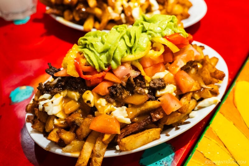 A plate of loaded fries inspired by Montreal food is topped with ground beef, diced tomatoes, cheese curds, guacamole, and sliced peppers on a colorful table. Another tempting plate of food is blurred in the background.