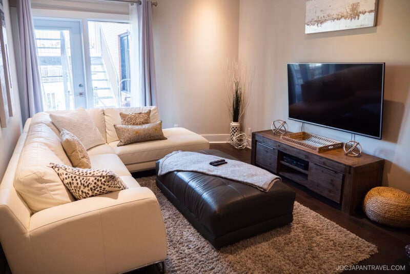 A modern Montreal living room with a white sectional sofa, patterned pillows, a dark brown ottoman, and a flat-screen TV on a wooden stand. Large windows let in natural light, and neutral decor accents the space.