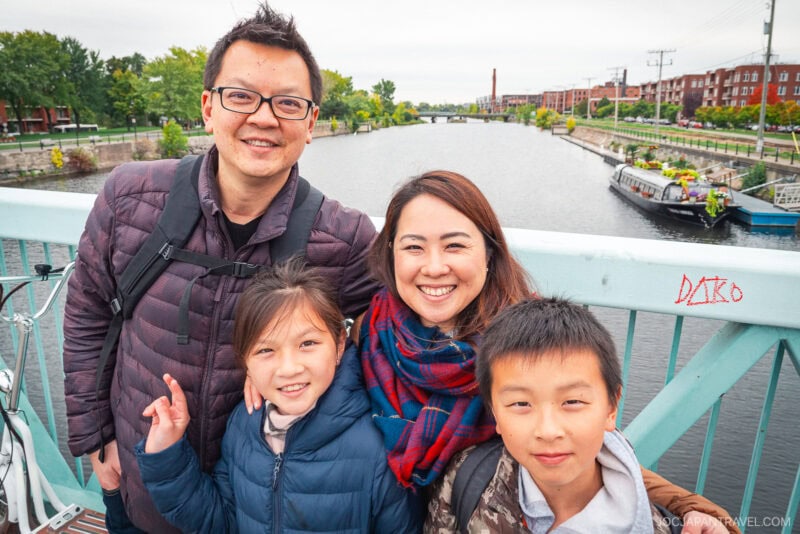 A smiling family of four, two adults and two children, poses on a bridge over a river in Montreal, with trees, boats, and red-brick buildings in the background on a cloudy day.