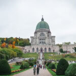 Montreal Itinerary for Families A large stone basilica with a green dome stands atop a hill in Montreal, surrounded by manicured gardens and trees with autumn foliage. People walk up a path toward the entrance, and cars are parked along the roads on either side.