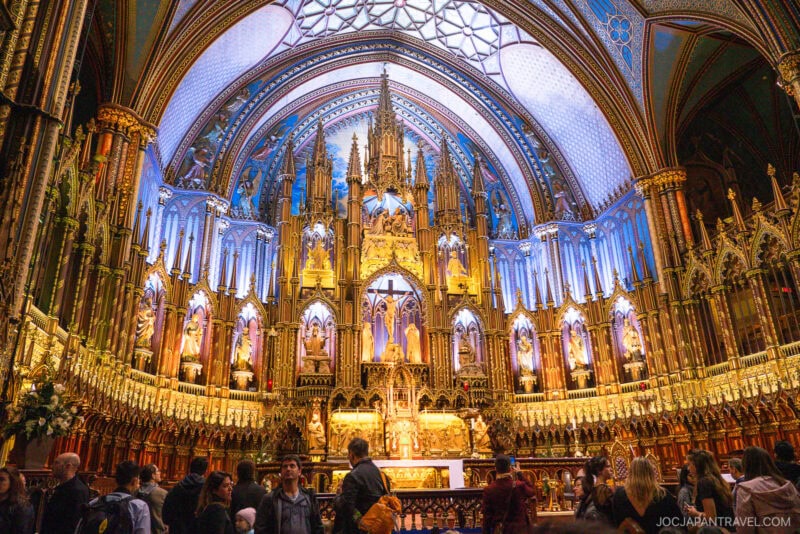 A group of people stands inside a grand, ornate Montreal church with a brightly illuminated, intricate altar featuring arches, statues, and gold details under a richly decorated blue ceiling.