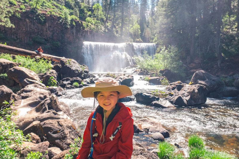 A smiling child in a wide-brimmed hat and red jacket sits on rocks near a flowing stream at Mount Shasta, with a waterfall and lush, sunlit forest behind. Another person appears on a fallen log above the water.
