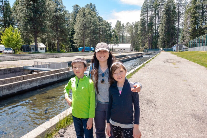 A woman and two children stand smiling on a sunny path near a long fish hatchery pool at the base of Mount Shasta, with tall trees and buildings in the background. The children wear bright jackets and the woman wears a hat and sunglasses.