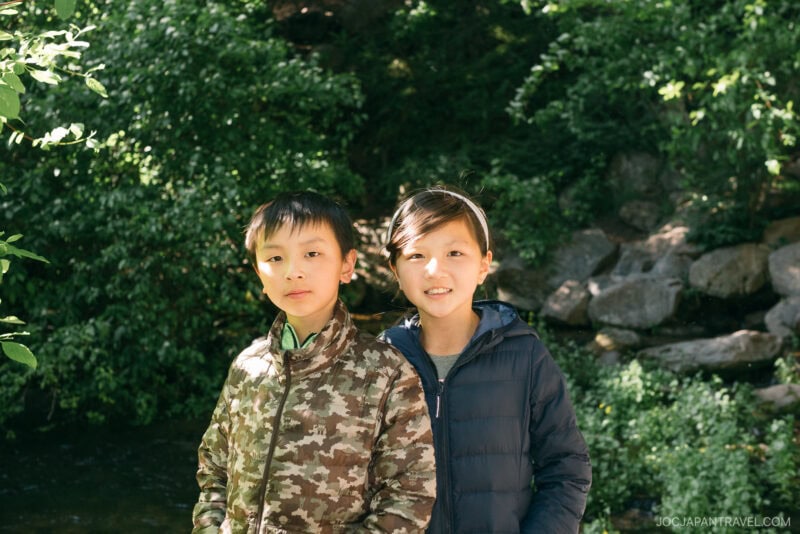 Two children pose outdoors in front of green foliage and rocks near Mount Shasta, wearing jackets. Sunlight filters through the trees, illuminating their faces.