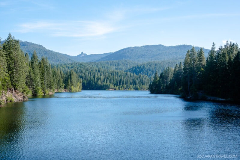 A calm blue lake, nestled beneath Mount Shasta, is surrounded by dense green pine trees and distant forested mountains under a clear blue sky.
