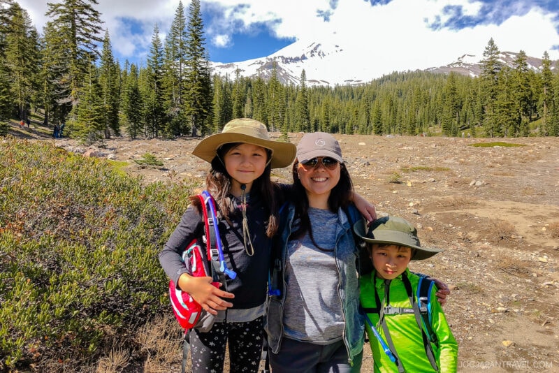 A woman and two children in hiking gear smile at the camera with pine trees and snow-capped Mount Shasta in the background on a sunny day.