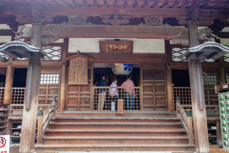A small group of people enters Myoryu-ji Temple, a traditional Japanese wooden temple with intricate carvings, sliding doors, and hanging lanterns above the entrance.