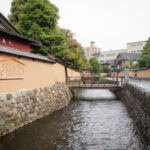 A narrow canal runs alongside traditional Japanese buildings in the Nagamachi Samurai District, with beige walls, stone foundations, and trees. A small wooden bridge crosses the canal as a few people walk on the quiet street nearby.