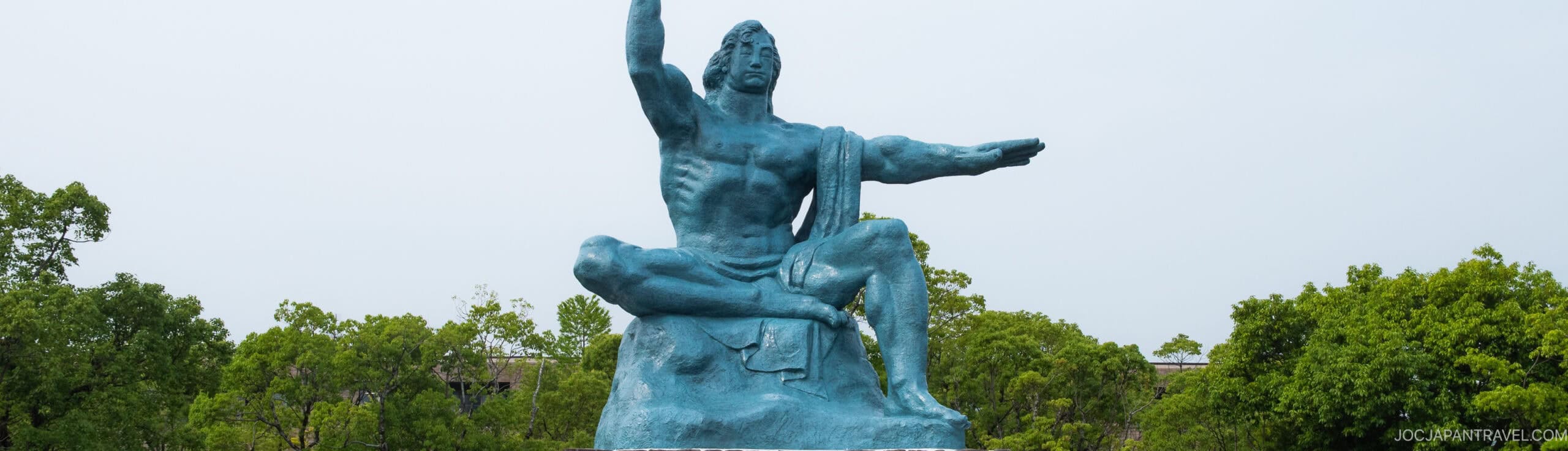 A large blue statue of a seated man with one arm raised and the other extended to the side, set against a background of green trees and a pale sky.