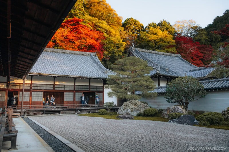 A traditional Japanese Zen garden with raked gravel, rocks, and manicured trees sits beside the wooden buildings of Nanzen-ji. Vibrant autumn foliage in red, orange, and yellow blankets the hillside in the background.
