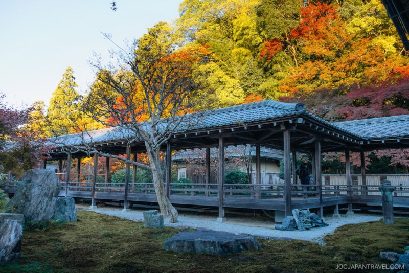 A traditional Japanese wooden walkway at Nanzen-ji stands amid autumn trees with vibrant red, yellow, and green leaves in a serene garden setting under clear blue skies.