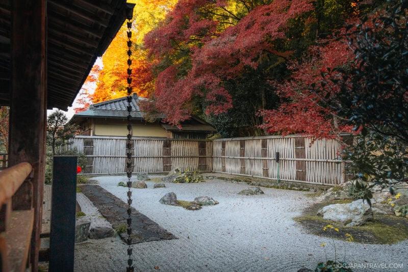 A tranquil Japanese rock garden at Nanzen-ji features white gravel, scattered rocks, autumn-colored trees, and a bamboo fence, all framed by traditional wooden buildings.
