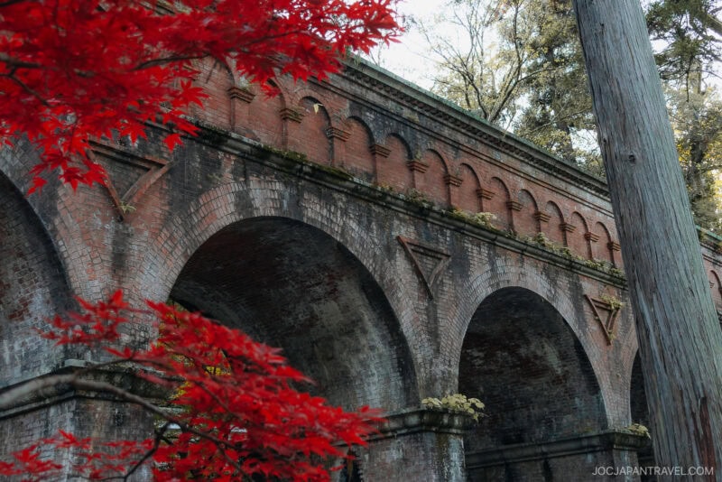 Historic brick aqueduct with large arches at Nanzen-ji, partially framed by vibrant red maple leaves in the foreground and a wooden utility pole to the right; trees are visible behind the structure.