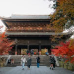 Tourists stand and take photos in front of the large, traditional wooden gate of Nanzen-ji Temple, surrounded by colorful autumn foliage in vibrant shades of red and orange.