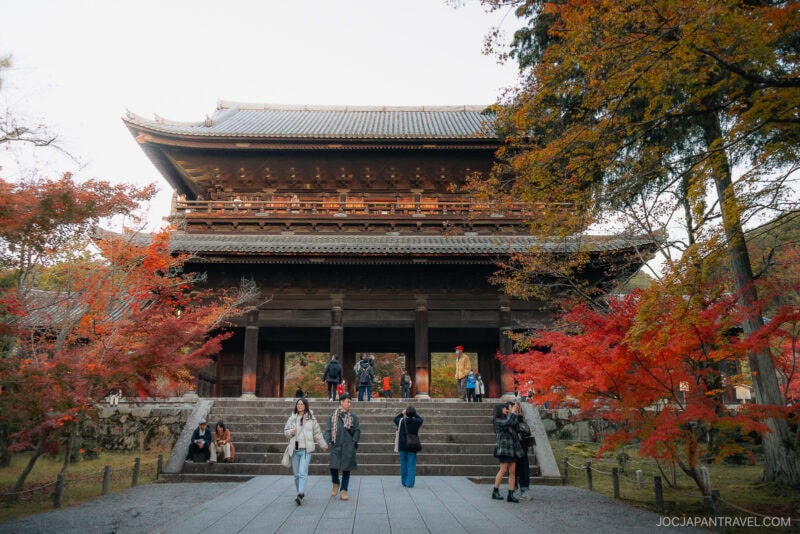 Tourists stand and take photos in front of the large, traditional wooden gate of Nanzen-ji Temple, surrounded by colorful autumn foliage in vibrant shades of red and orange.