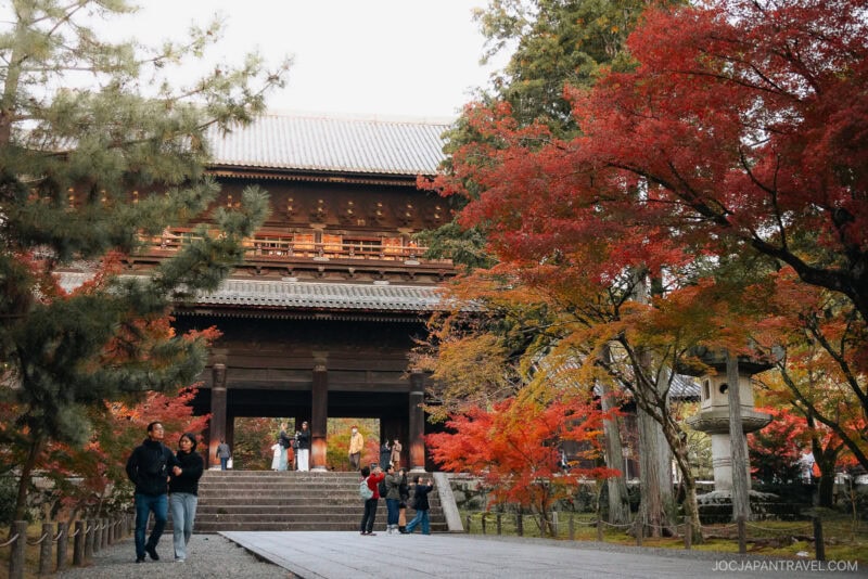 A group of people walk and take photos near the large wooden Nanzen-ji temple gate, surrounded by vibrant autumn trees with red, orange, and yellow leaves. Stone lanterns line the path.