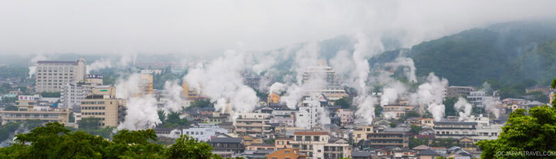 A panoramic view of a city with numerous buildings and steam rising from multiple spots, set against green, misty hills in the background. The atmosphere appears humid and overcast.