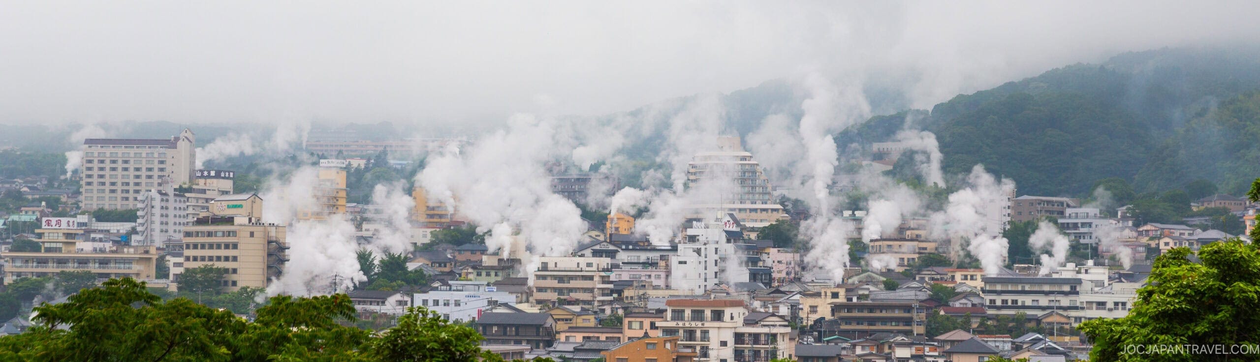A panoramic view of a city with numerous buildings and steam rising from multiple spots, set against green, misty hills in the background. The atmosphere appears humid and overcast.