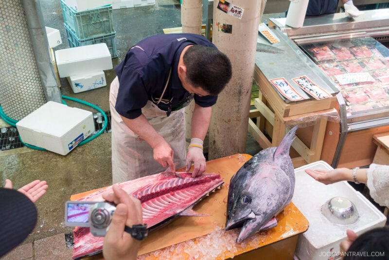 A fishmonger fillets a large tuna on a table at Omicho Market, with the fish cut open to reveal its bones and flesh. Several people watch and take photos, and packaged fish products are seen nearby.
