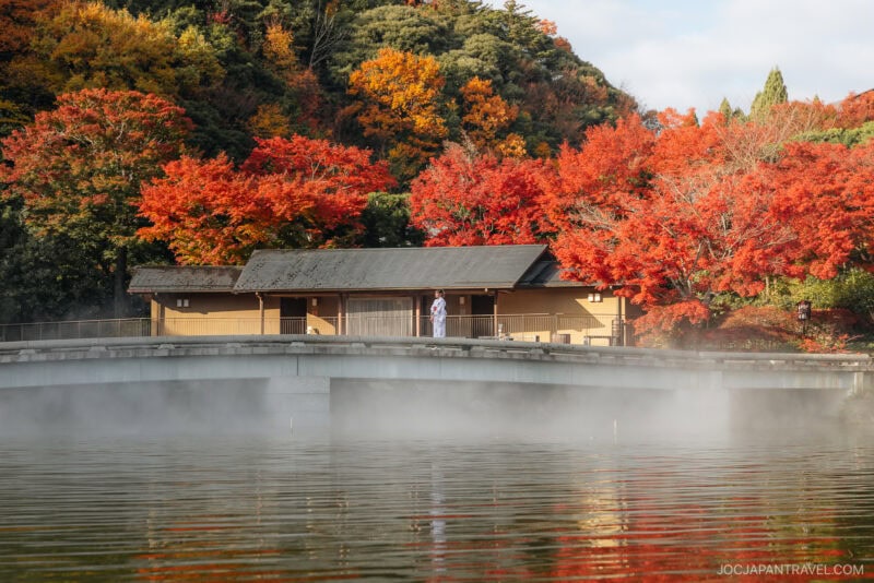 A person stands on a stone bridge over water, surrounded by vibrant red and orange Osaka autumn foliage, with mist rising from the water and a traditional Japanese building in the background.