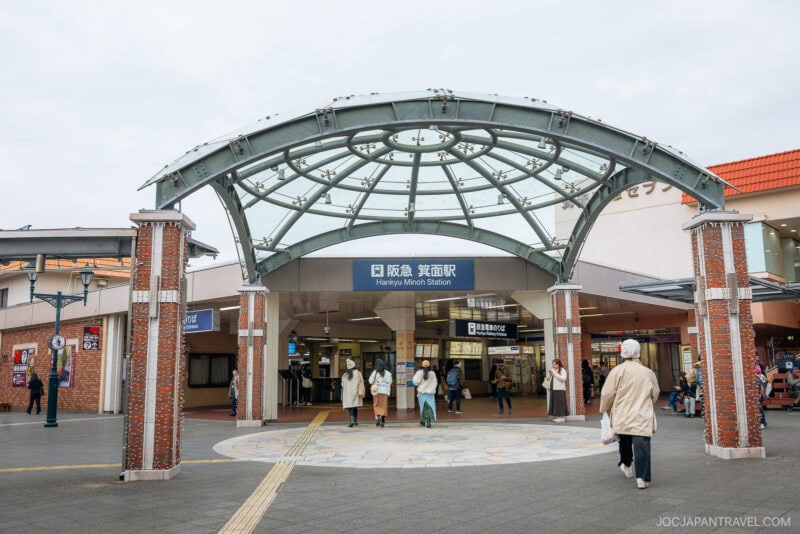 People walk near the entrance of Hankyu Minoh Station in Japan, which features a glass and metal canopy supported by brick columns. The station is a popular starting point for Osaka autumn foliage excursions. The station sign is visible above the entrance.