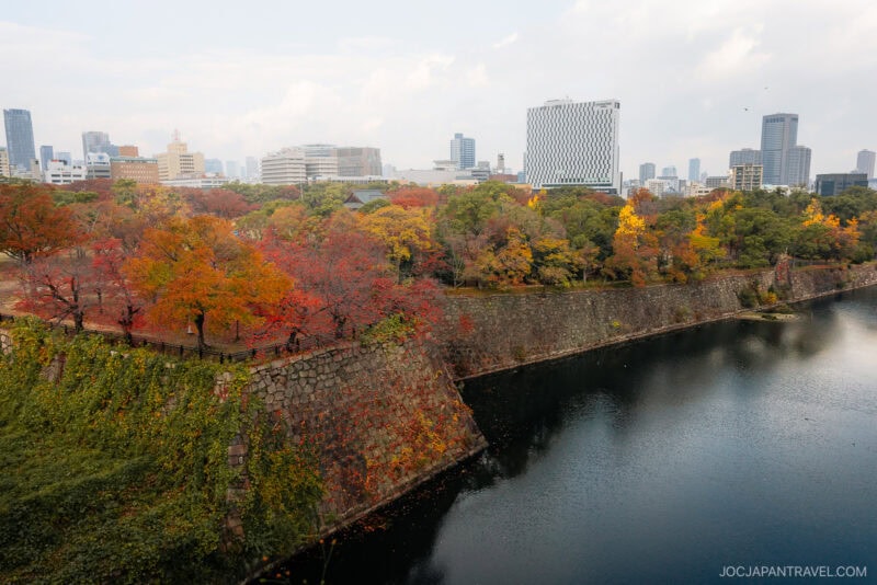 Cityscape with modern buildings in the background, Osaka autumn foliage of red and yellow leaves lining a stone wall along a river or moat under a partly cloudy sky.