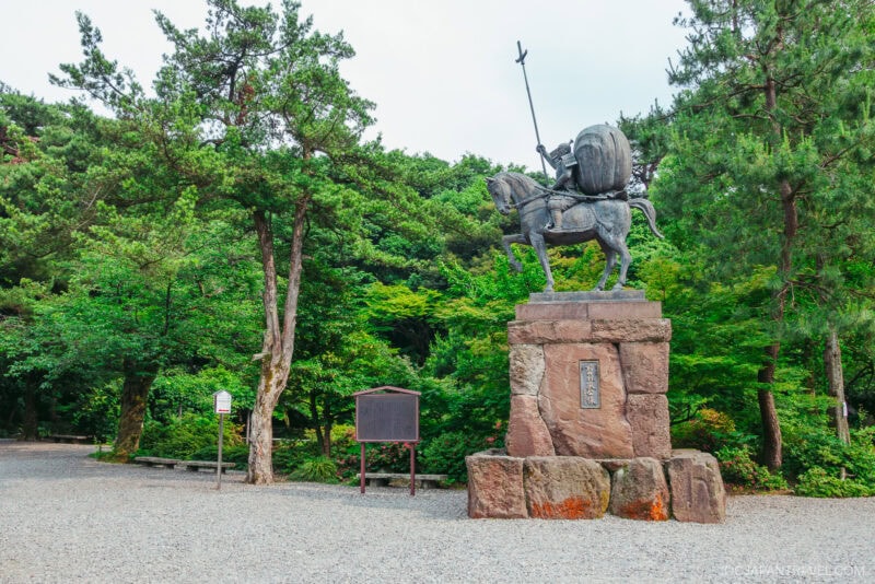 A bronze statue of a warrior on horseback holding a spear stands on a stone pedestal, surrounded by trees and greenery in the peaceful grounds of Oyama Shrine.