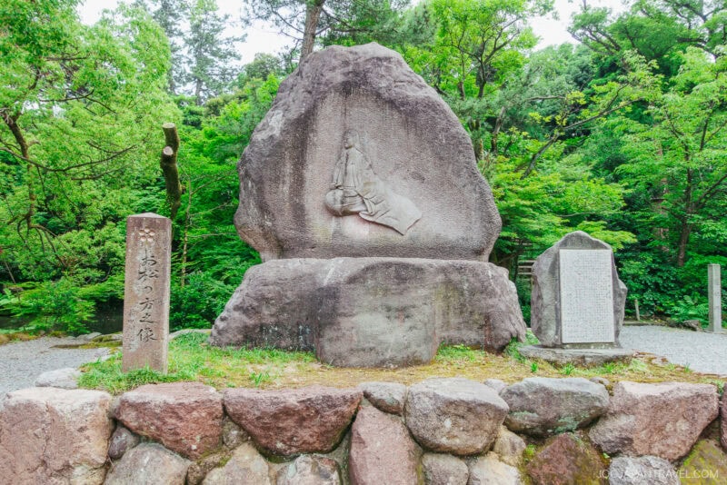 A large stone monument at Oyama Shrine features a carved relief of a reclining figure, surrounded by lush green trees, with two vertical stone markers and a plaque nearby. The monument sits atop a stone platform.