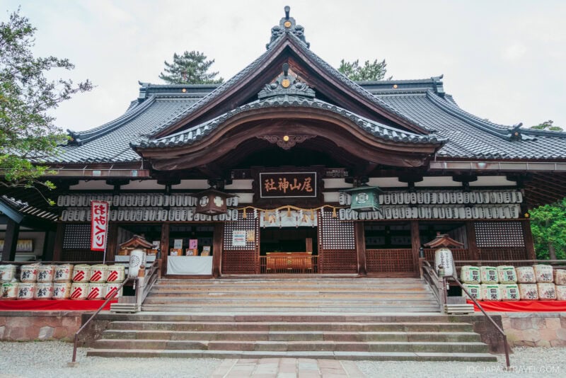 A traditional Japanese shrine, Oyama Shrine features a tiled roof, ornate wooden carvings, hanging lanterns, and stone steps leading to the entrance. Barrels are stacked on either side of the steps and trees frame the building.