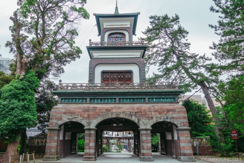 A historic Japanese gate at Oyama Shrine with three tiers, arched entrances, and a clock tower stands among tall trees and greenery on a cloudy day.