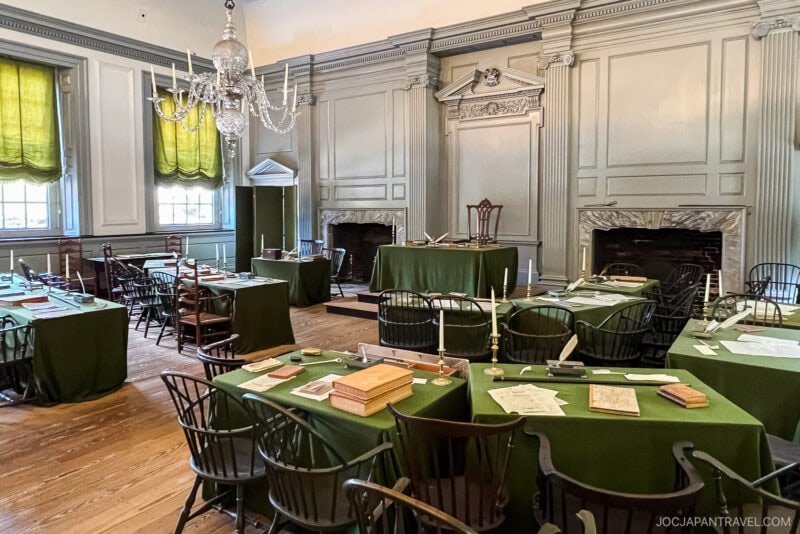 A historic Philadelphia room with green-covered tables, wooden chairs, and scattered papers, books, and quills. Large windows with green drapes, ornate walls, and a chandelier create a formal, colonial-era meeting atmosphere.